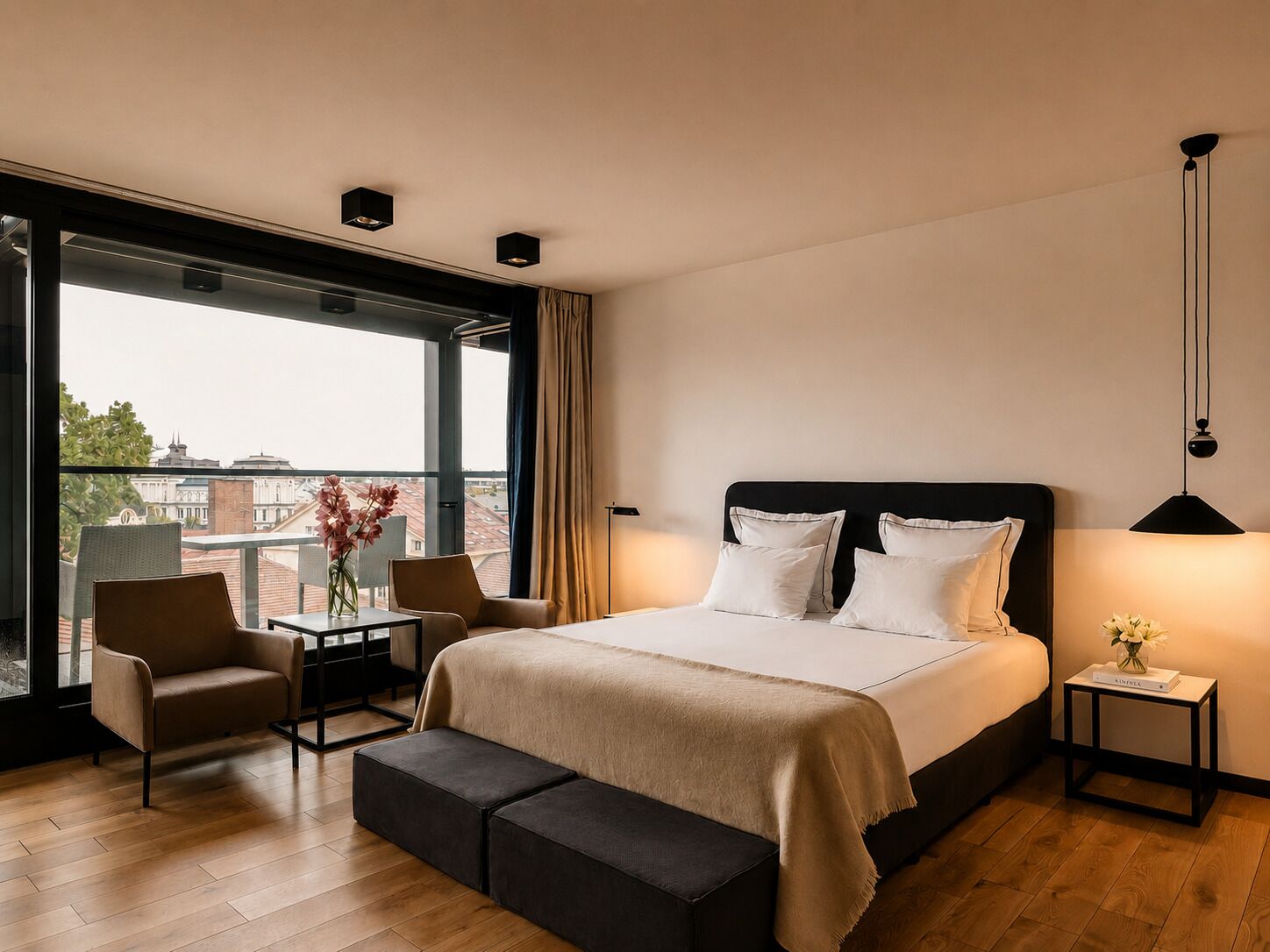 Habitación de hotel moderna con una cama grande, una mesita, dos sillas, un banco y vistas a la ciudad a través de amplios ventanales.