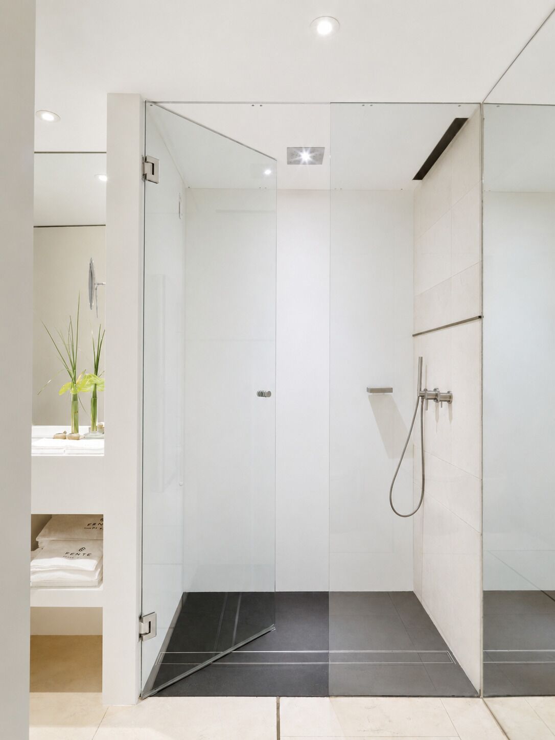 Un baño moderno con cabina de ducha de vidrio, suelo de baldosas blancas y una planta pequeña a la izquierda.