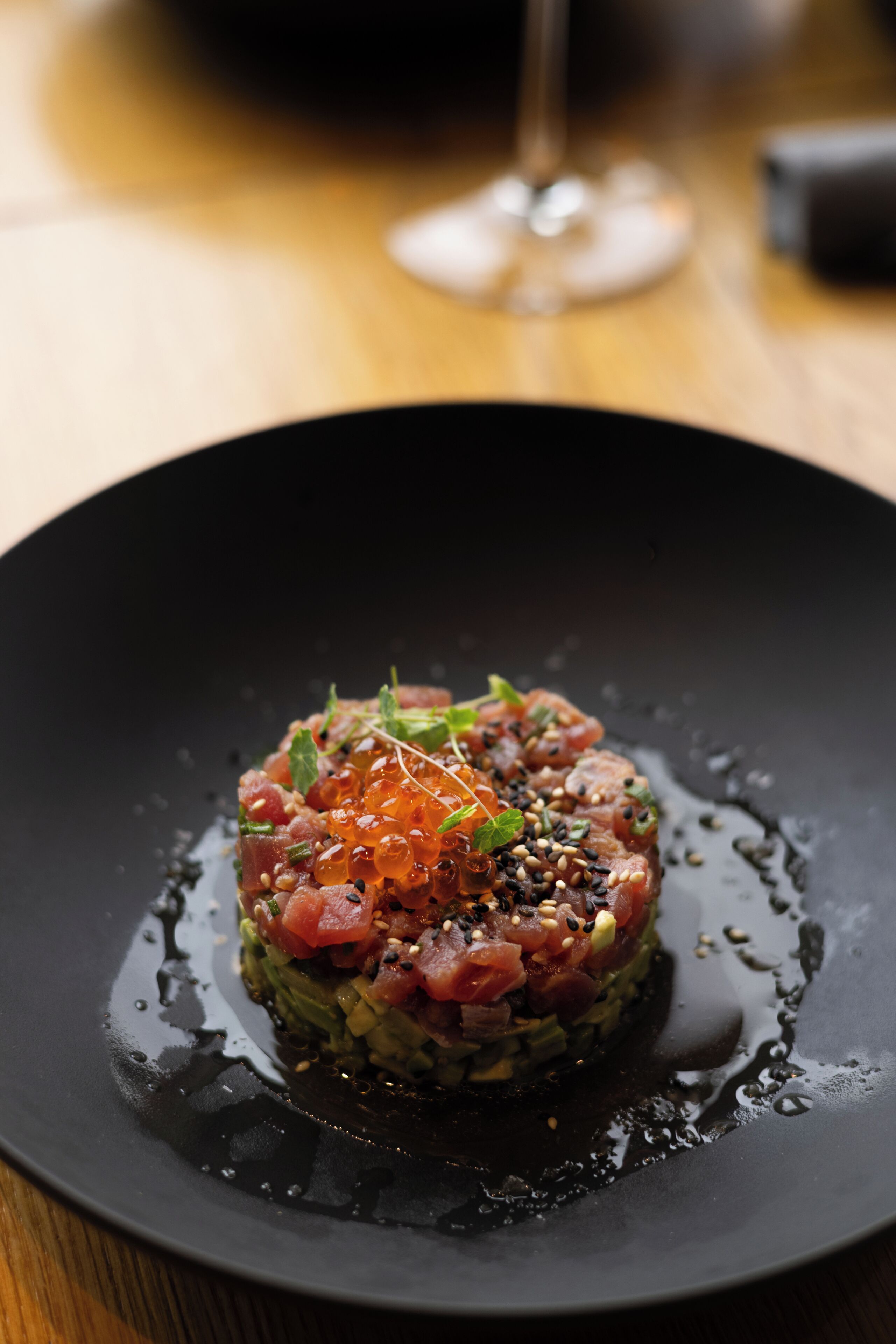 A black plate with a dish of raw fish salad, garnished with sesame seeds and herbs, on a wooden table.
