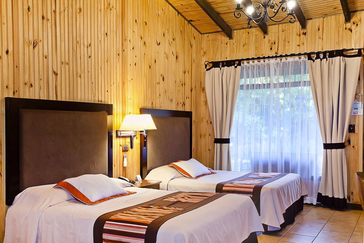 Um quarto com painéis de madeira, duas camas, um candelabro e uma janela com cortinas.
