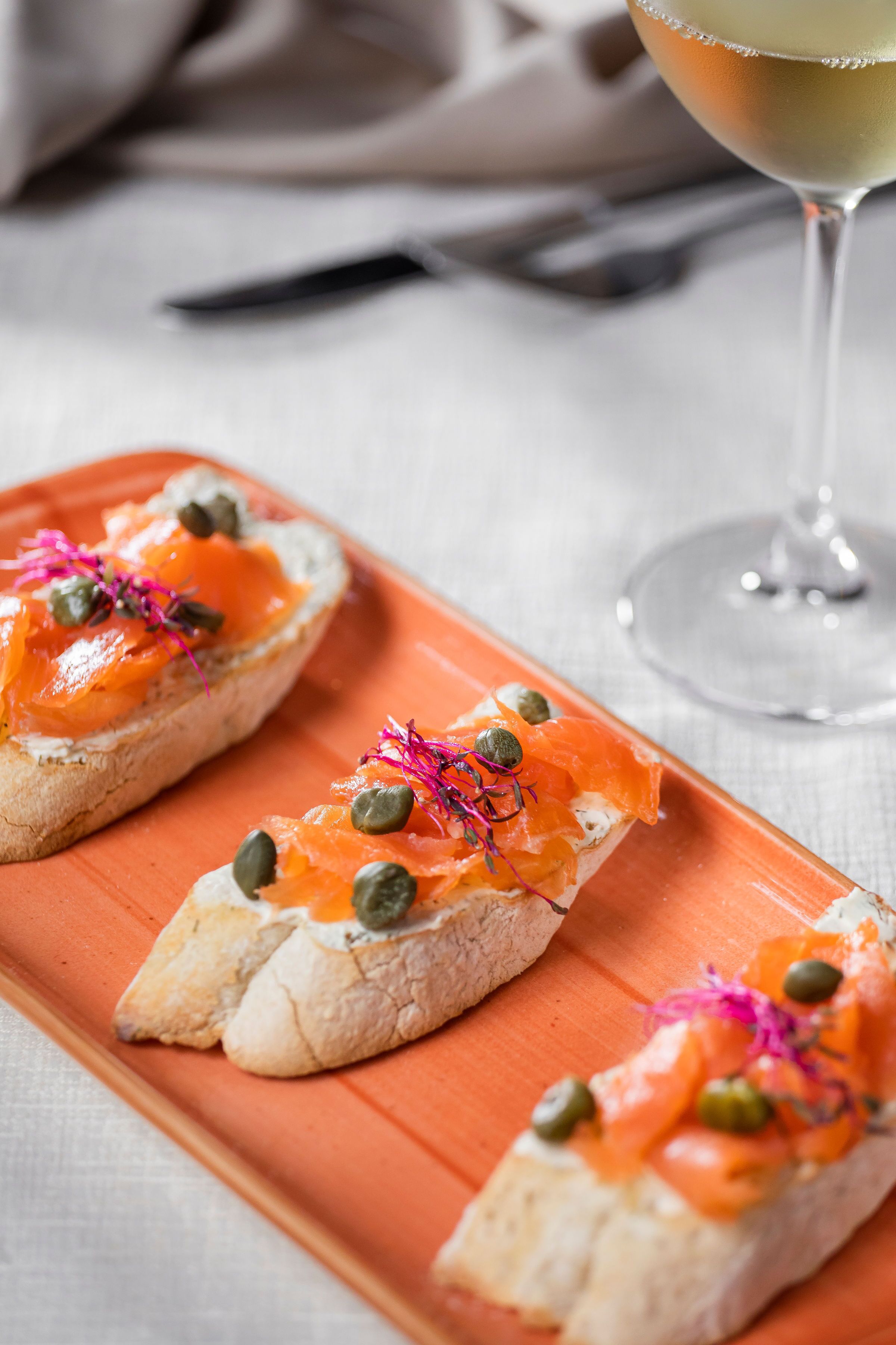 Three open-faced sandwiches with smoked salmon, capers, and pink edible flowers on a rectangular orange plate.