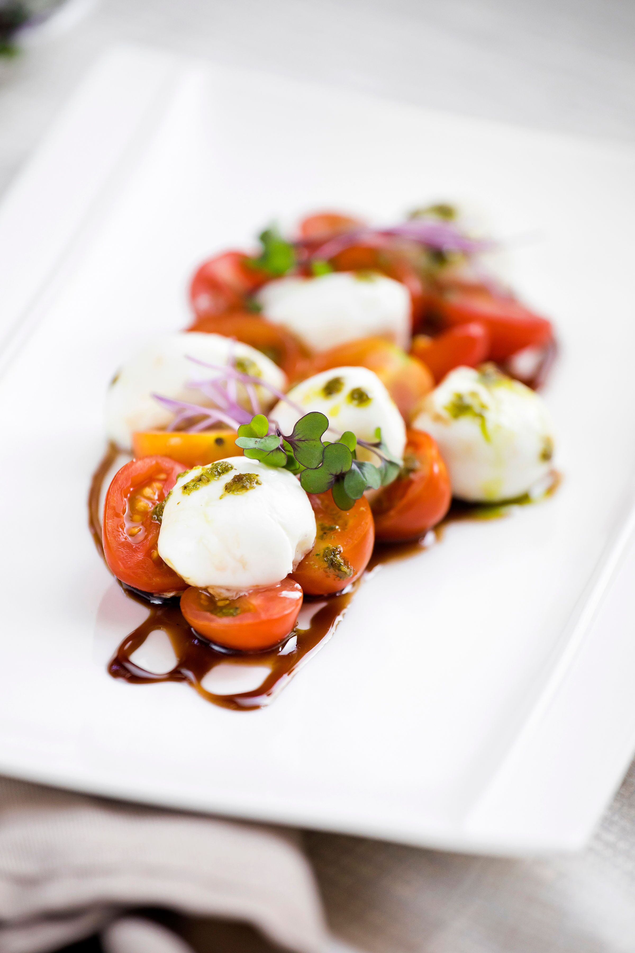 A white rectangular plate with a food arrangement consisting of cherry tomatoes, mozzarella balls, and a drizzle of sauce.