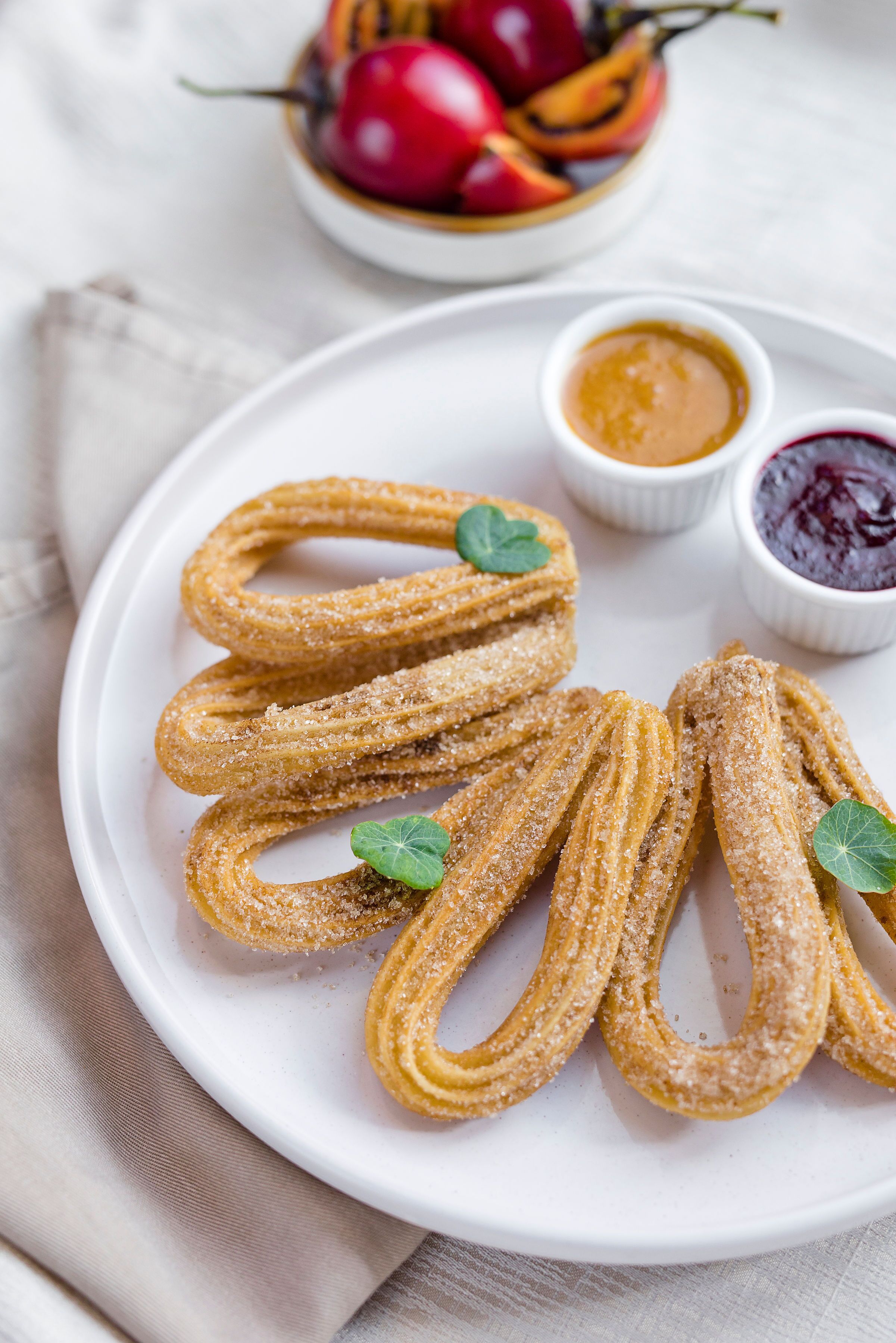 Ein Teller mit Churros, Zucker und zwei Dip-Saucen, im Hintergrund eine Fruchtbowl.