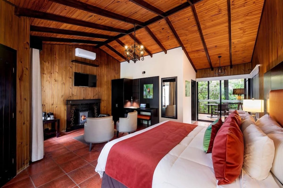A room with a wooden ceiling, a fireplace, a bed with pillows, a desk with a chair, and a television.