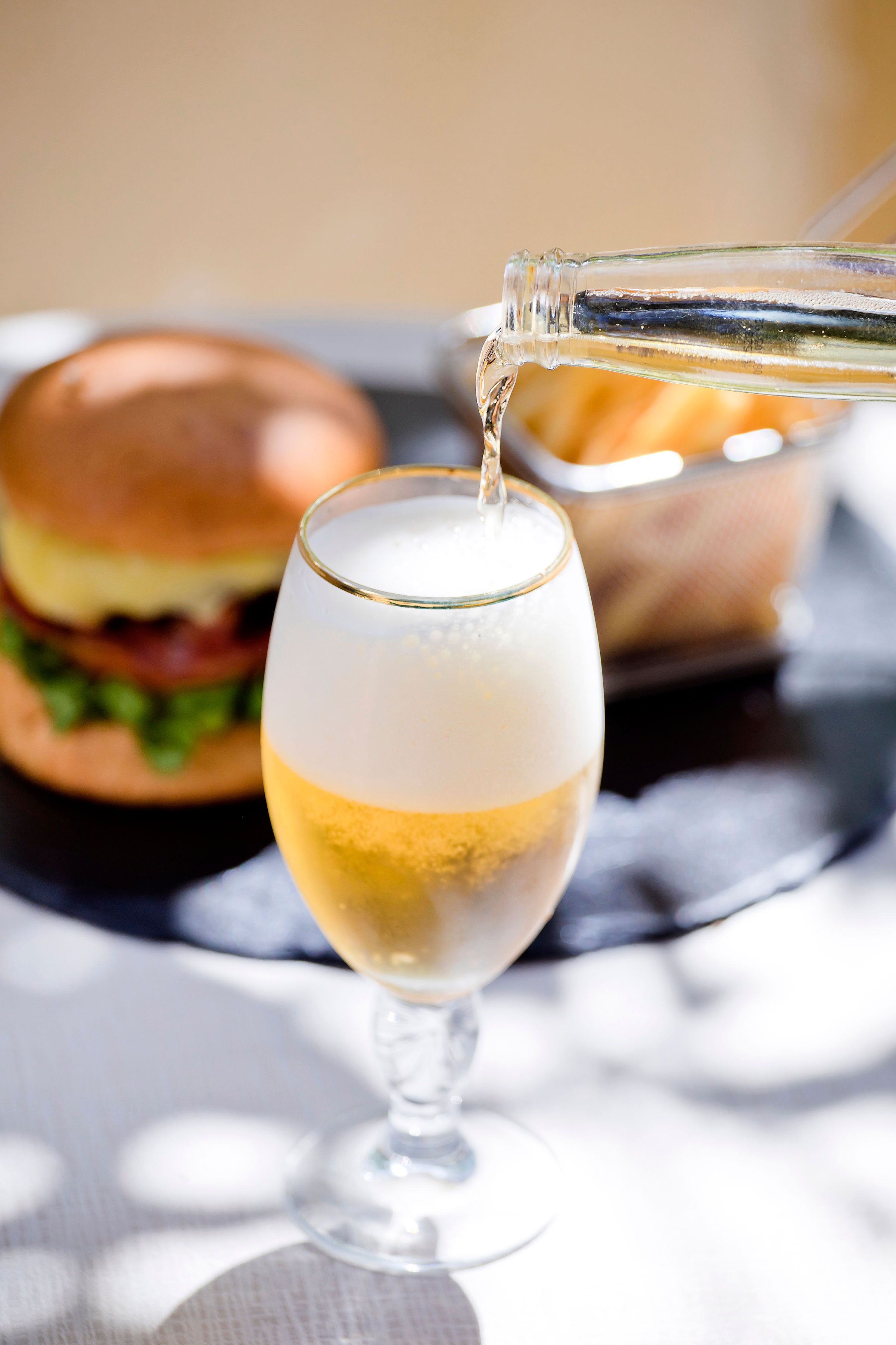 A glass of white wine being poured into a stemmed glass. In the background, there is a burger on a plate.