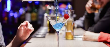 A bar setting with a martini glass on the counter, a cocktail shaker pouring a blue liquid, and a cherry garnish.