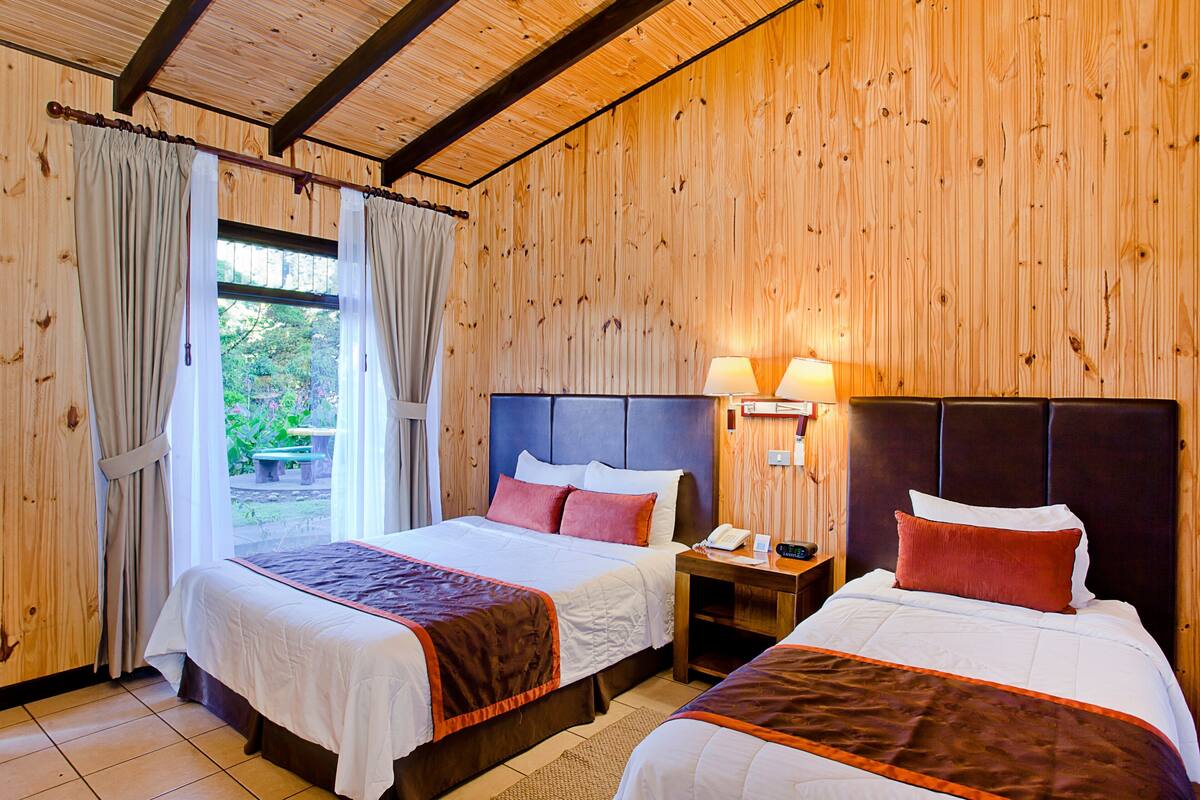Quarto com duas camas, paredes de madeira, janela com cortinas e mesa de cabeceira com abajur.