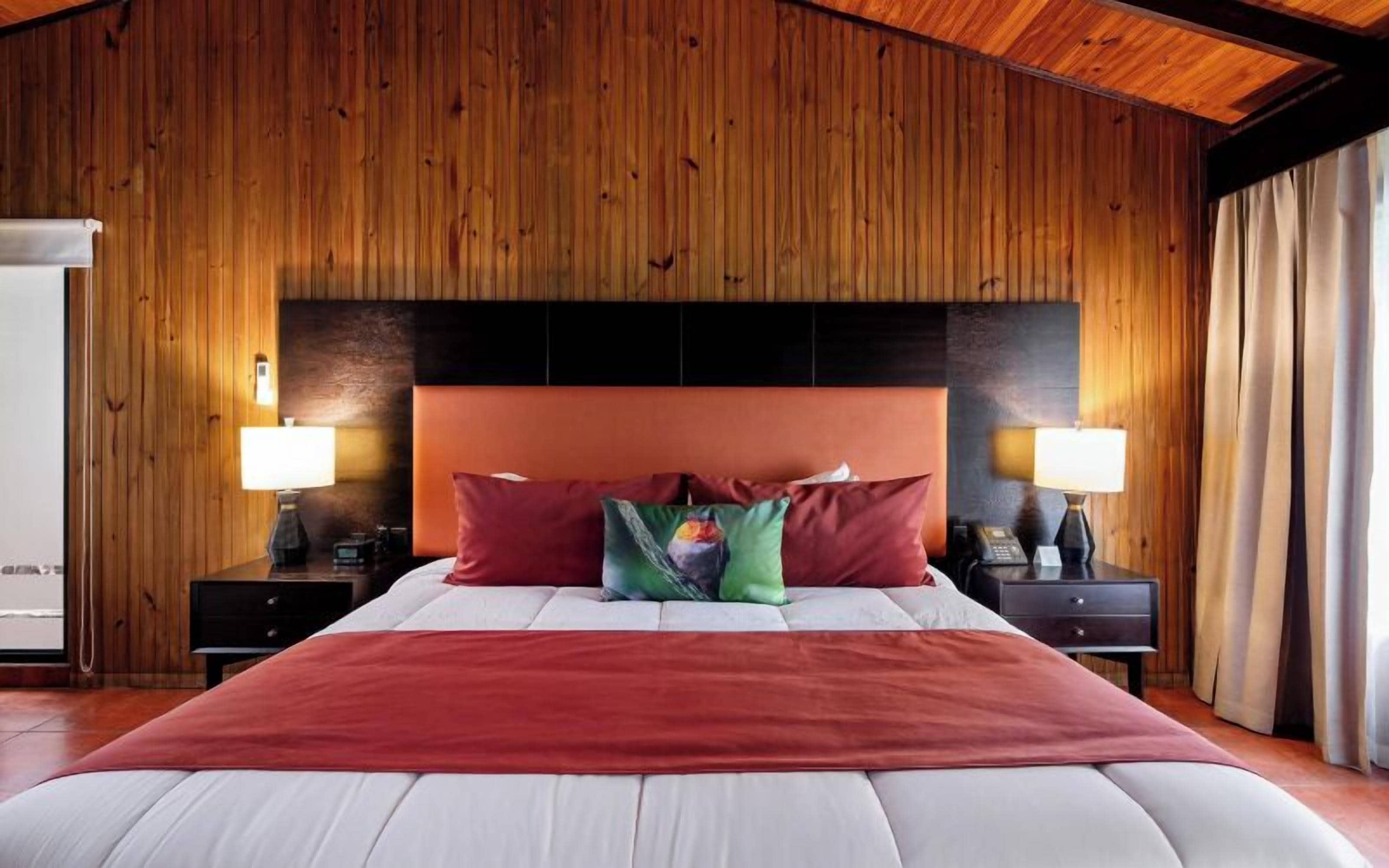 Habitación de hotel con pared de paneles de madera, una cama grande con almohadas rojas y verdes, dos mesitas de noche con lámparas y vistas al exterior.