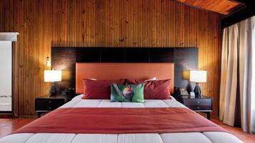 Habitación de hotel con pared de paneles de madera, una cama grande con almohadas rojas y verdes, dos mesitas de noche con lámparas y vistas al exterior.