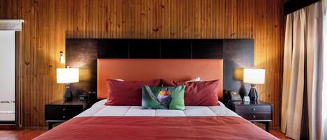 Une chambre d’hôtel avec un mur lambrissé en bois, un grand lit avec des oreillers rouges et verts, deux tables de chevet avec des lampes, et une vue sur l’extérieur.