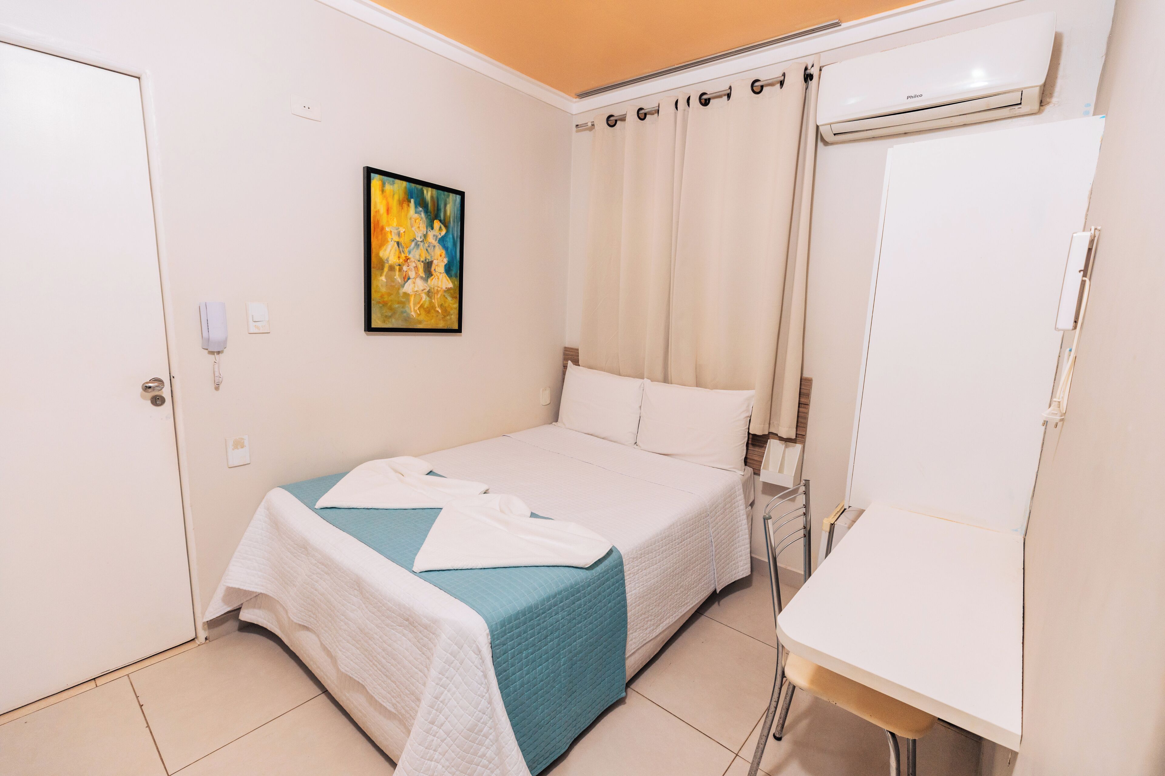 Ein Hotelzimmer mit Bett, Schreibtisch, Stuhl, einem Gemälde und einem Vorhang.