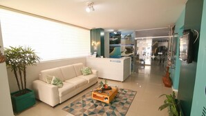 Un salón moderno con sofá, mesa de centro y plantas en macetas. Se observa un mostrador de recepción al fondo.
