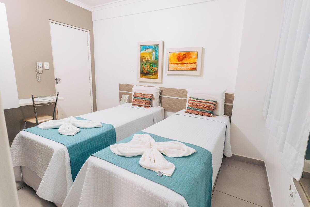 A hotel room with two beds, each with a teal bedspread and white towels. There are two framed paintings on the wall, a white door, and a chair in the corner.