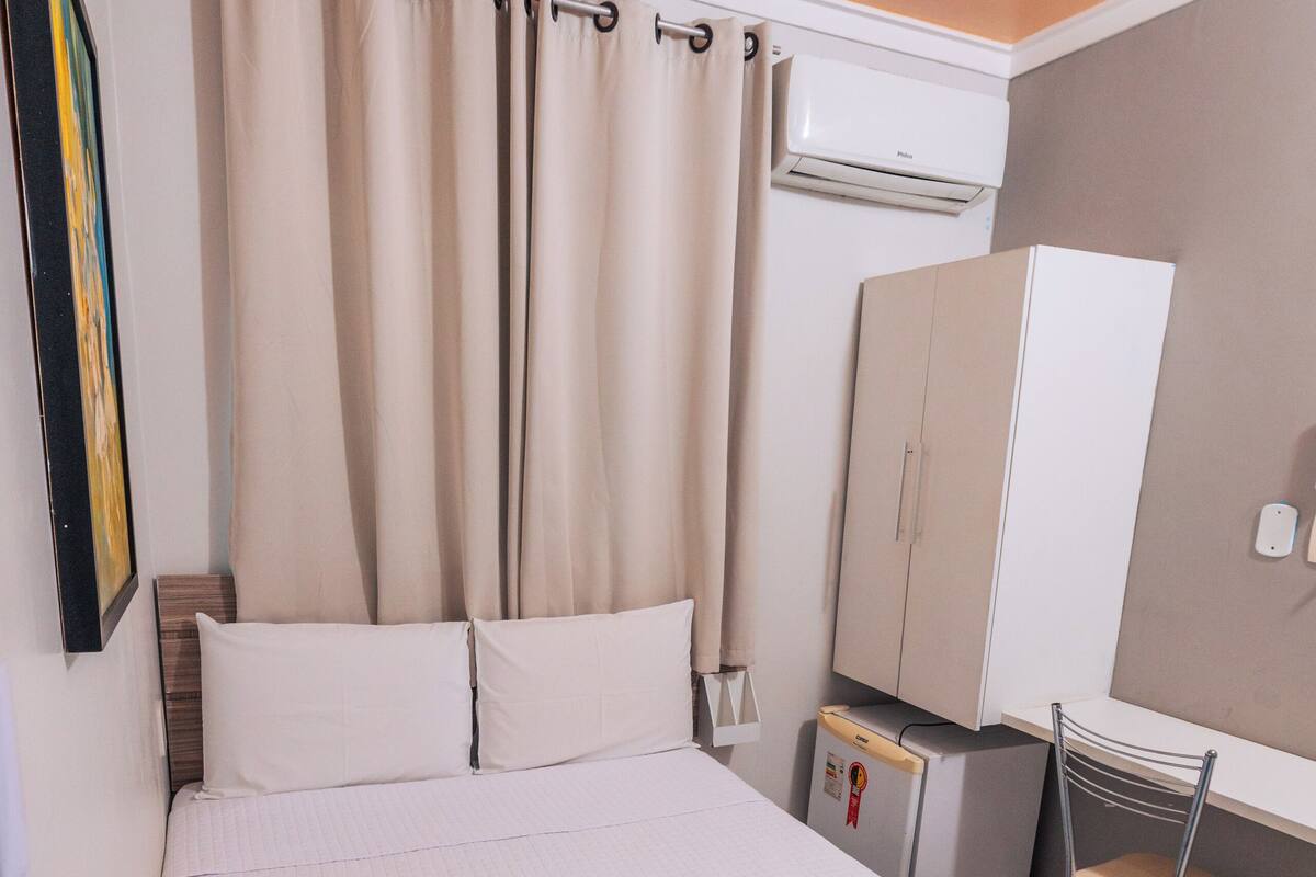 A hotel room with a bed, a desk, a chair, an air conditioning unit, and a wardrobe.