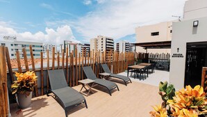 Terraza en la azotea con tumbonas, una mesa y sillas, rodeada por una valla de madera y plantas en macetas.