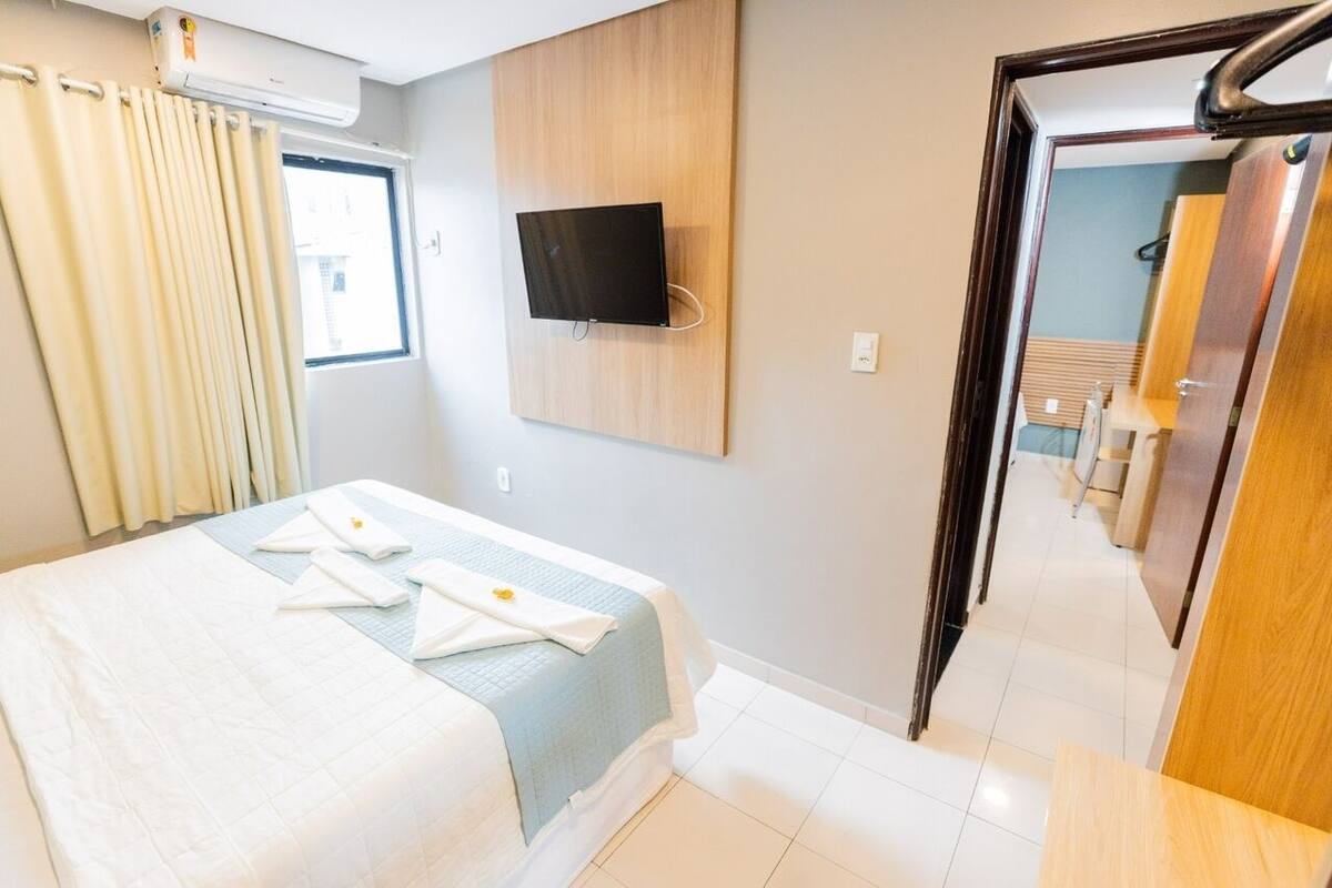 A hotel room with a bed, a TV mounted on the wall, a window with curtains, and a bathroom visible through an open door.