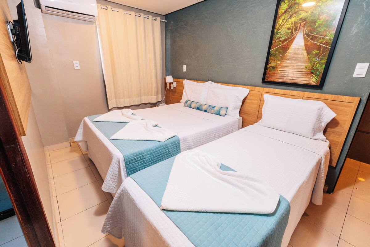 A hotel room with two beds, a painting of a forest bridge, and a window with curtains.