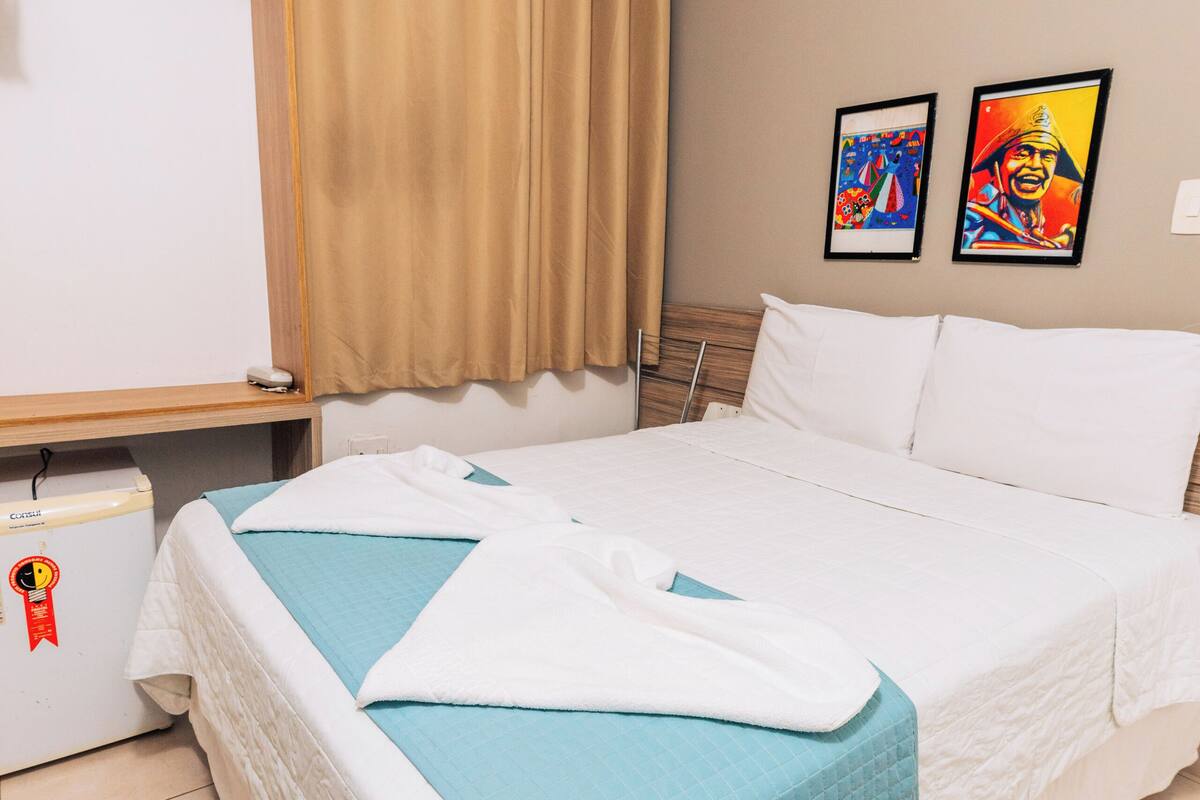 A hotel room with a bed, a nightstand, a heater, and two paintings on the wall.