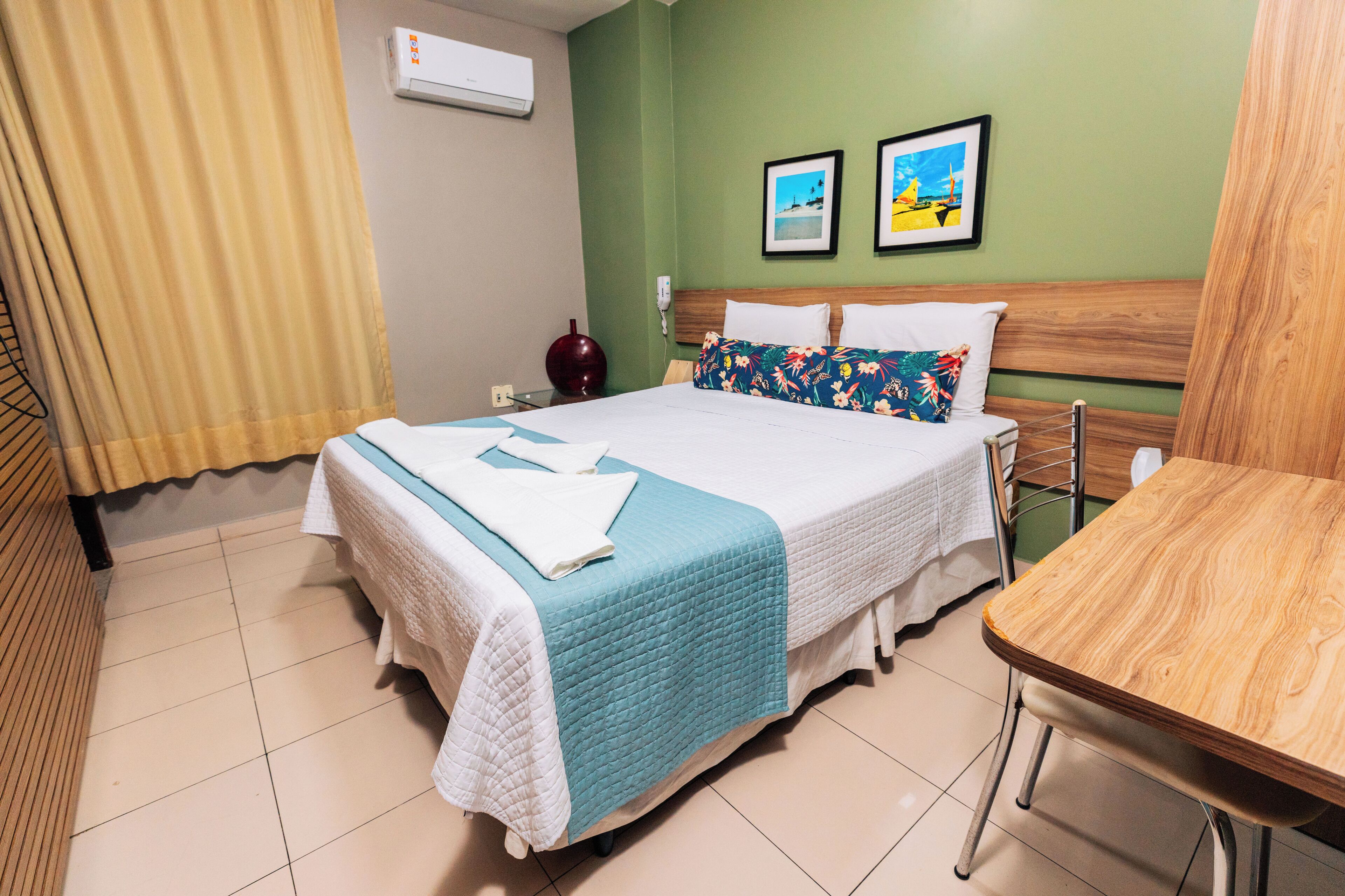 A hotel room with a bed, a wooden table, and two framed pictures on the wall.