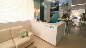 Un área de recepción con un mostrador blanco, un sofá color crema con un cojín verde y un muro de vidrio con un mural tropical.