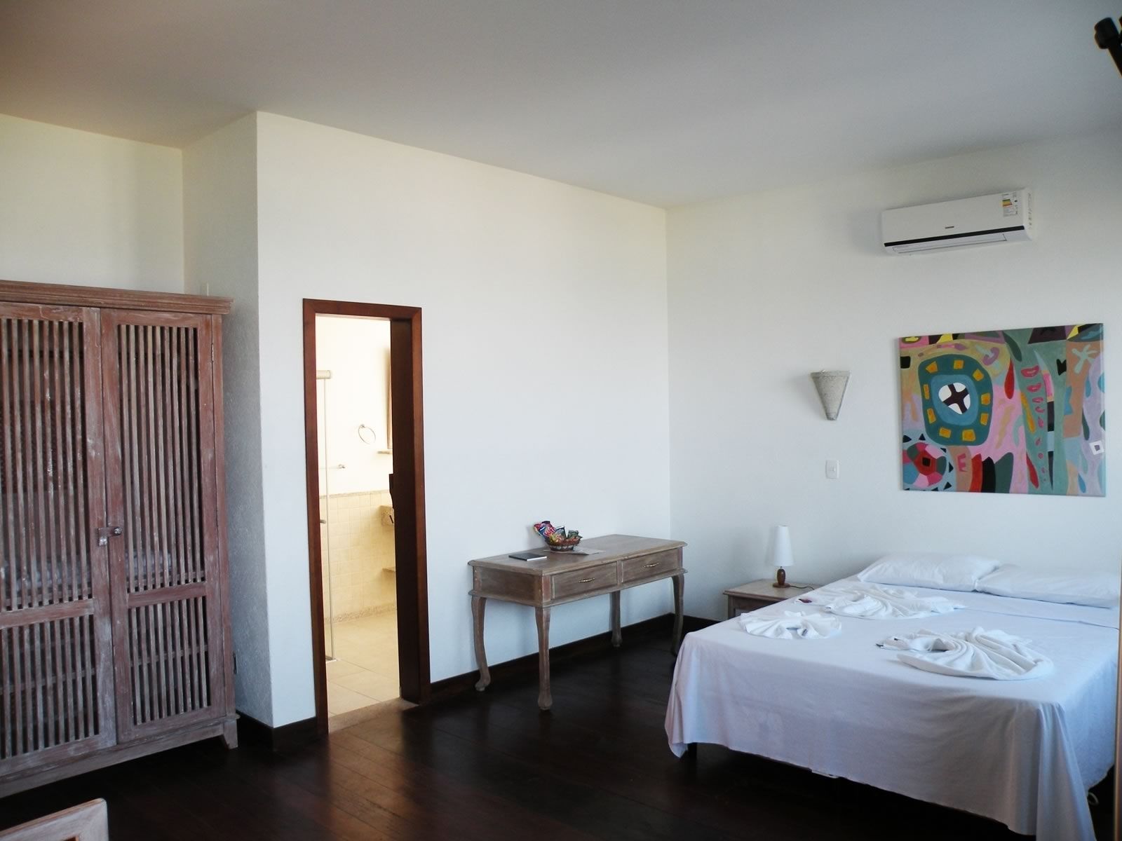 Una habitación de hotel con una cama, un armario de madera, una mesita con un jarrón, un cuadro en la pared y un equipo de aire acondicionado.