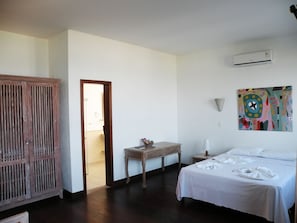 Una habitación de hotel con una cama, un armario de madera, una mesita con un jarrón, un cuadro en la pared y un equipo de aire acondicionado.