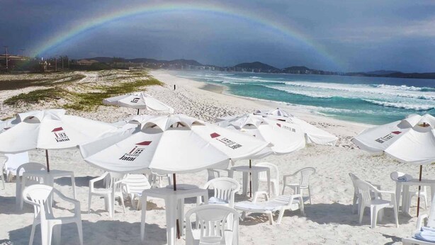 Une plage avec des parasols et des chaises, un arc-en-ciel, et une vue sur l’océan et les collines.