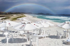 Una playa con sombrillas y sillas, un arcoíris y vistas al océano y las colinas.