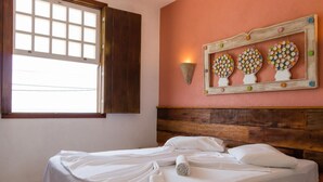 Une chambre avec une tête de lit en bois, un lit recouvert de draps blancs, une fenêtre avec des volets ouverts et un luminaire mural orné de motifs floraux.