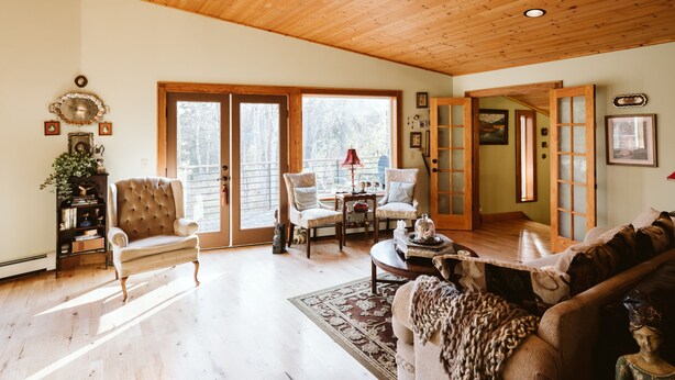 A spacious living room with a high wooden ceiling, large sliding glass doors leading to a deck, and a cozy fireplace area. - Maria's Creekside B&B (Anchorage)