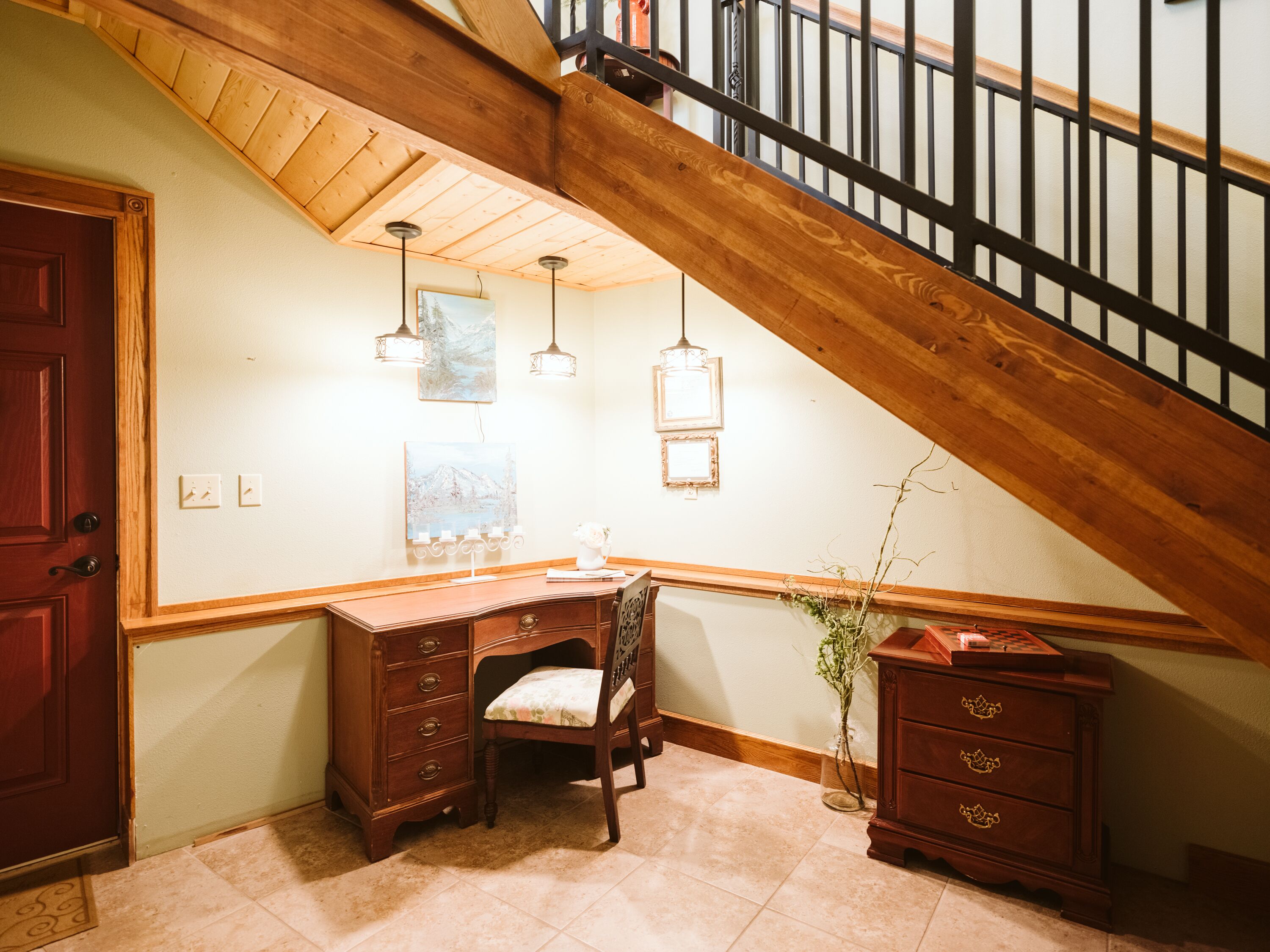 A corner room with a wooden desk, chair, and dresser. There is a staircase with a wooden railing and a door on the left.
