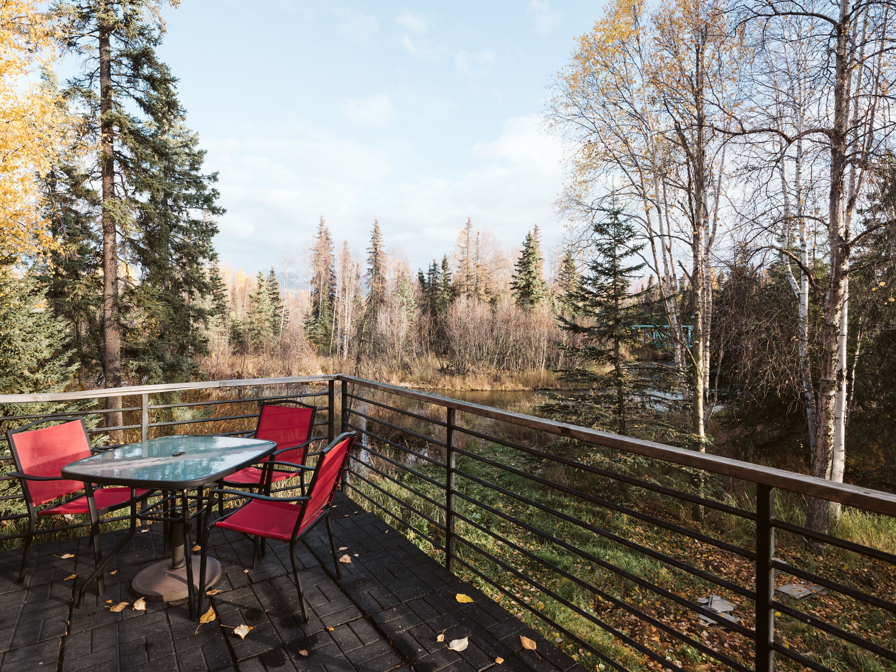 An outdoor deck with a glass table and red chairs, overlooking a forested area and a body of water.