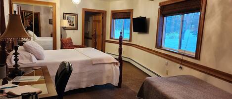 Un dormitorio con cama, mesita de noche, lámpara, silla y ventanas.