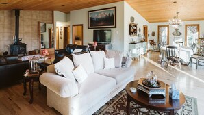 A spacious living room with a large sofa, a fireplace, a television, and a dining area with a chandelier. - Maria's Creekside B&B (Anchorage)