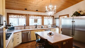 A kitchen with wooden ceiling, a chandelier, a stainless steel refrigerator, and a dining area with chairs. - Maria's Creekside B&B (Anchorage)