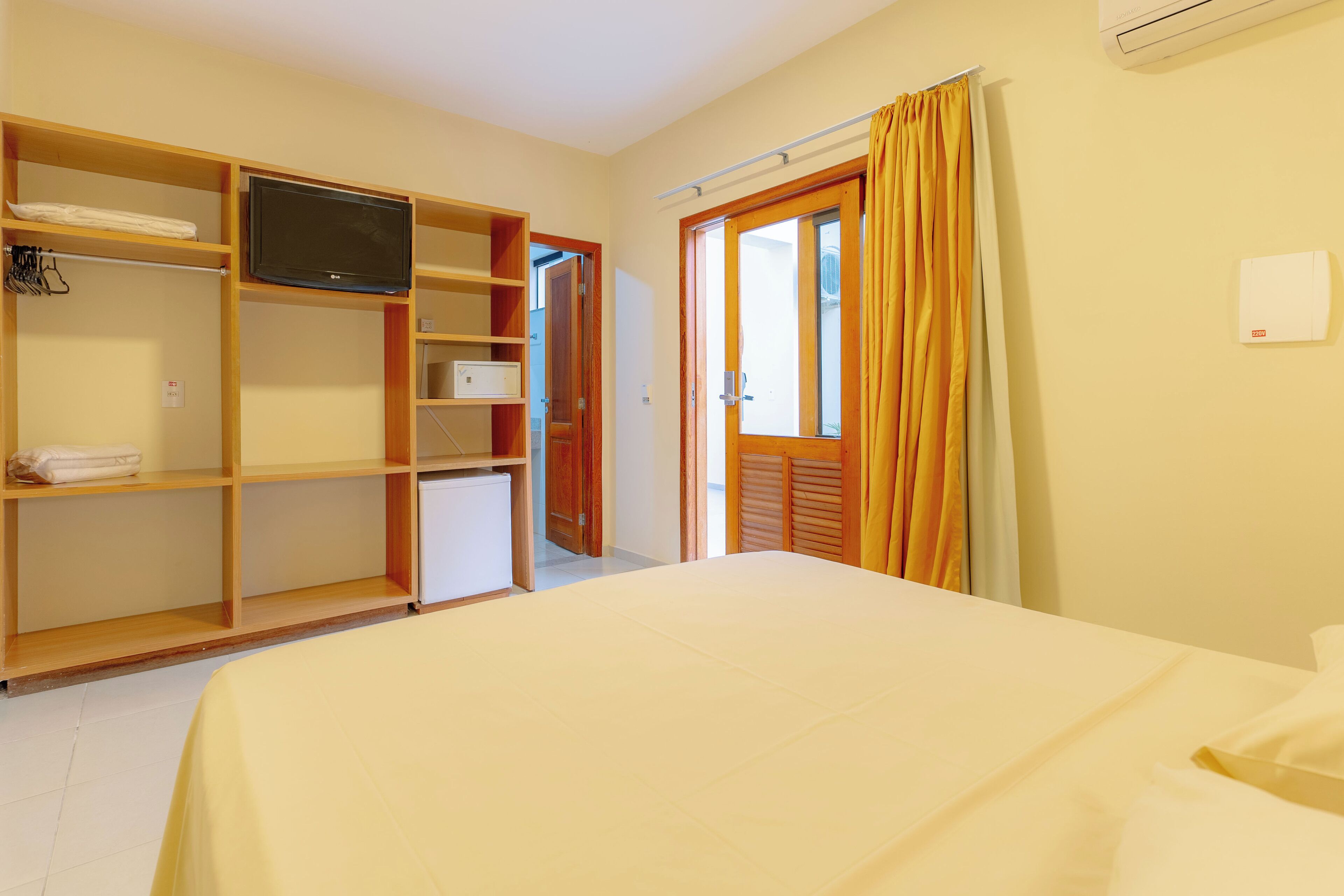 Quarto com cama, televisão em prateleira, guarda-roupa e porta para o exterior.