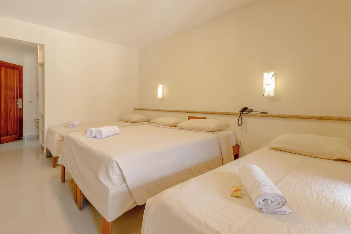 A hotel room with three single beds, each with a rolled towel and a small item on the bed. There are two wall-mounted light fixtures and a telephone on the wall.