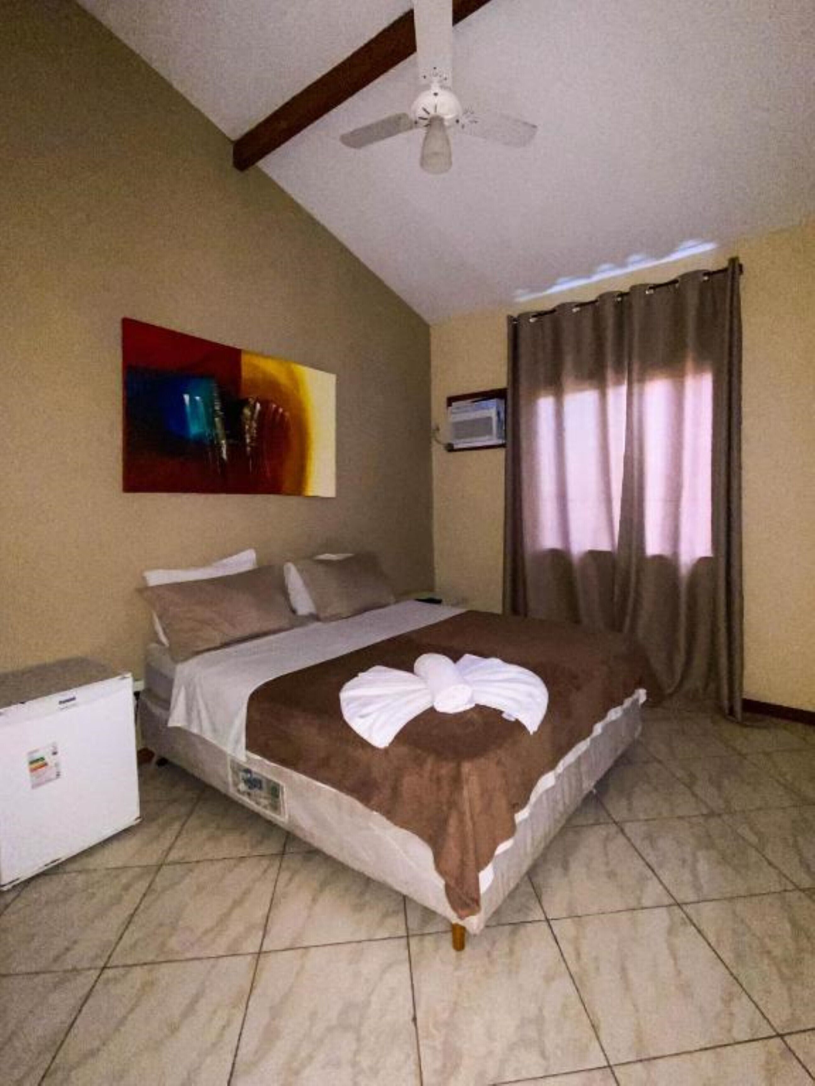 Quarto de hotel com cama, ventoinha no teto, quadro na parede, janela com cortinas e um pequeno frigorífico.