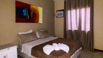 Quarto de hotel com cama, ventoinha no teto, quadro na parede, janela com cortinas e um pequeno frigorífico.