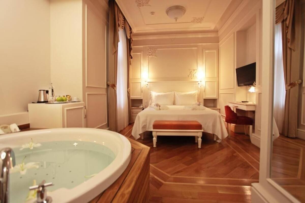 A hotel room with a large bathtub, a bed with white linens, a desk with a chair, and a television on the wall.