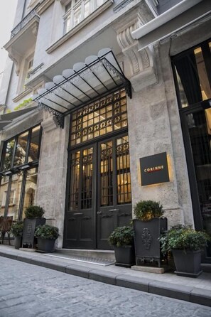Un bâtiment historique avec une porte d’entrée noire, une enseigne portant le nom « CORINNE » et des plantes en pot disposées sur le trottoir.