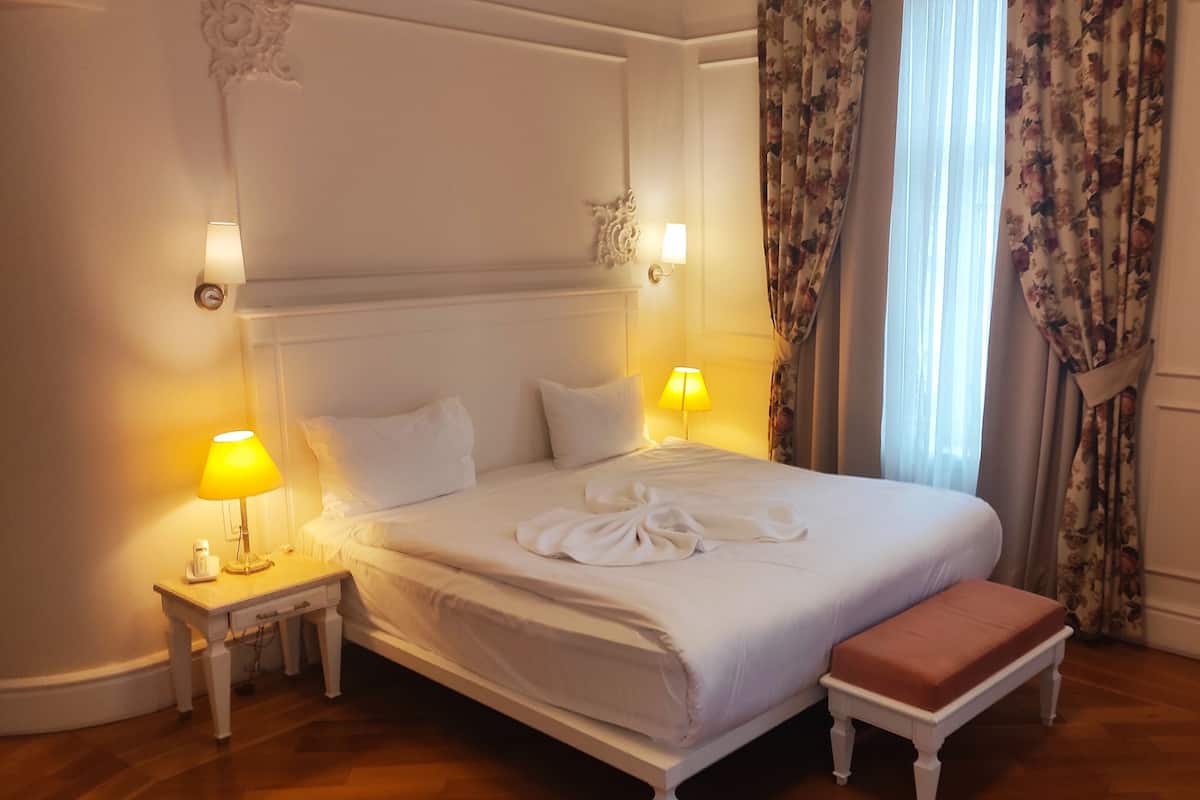 A neatly made bed with white linens, two bedside lamps, a floral curtain, and a wooden bench.