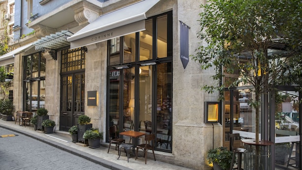 Un bâtiment doté d’une façade en pierre, de grandes fenêtres et d’un espace extérieur aménagé avec des tables et des chaises.