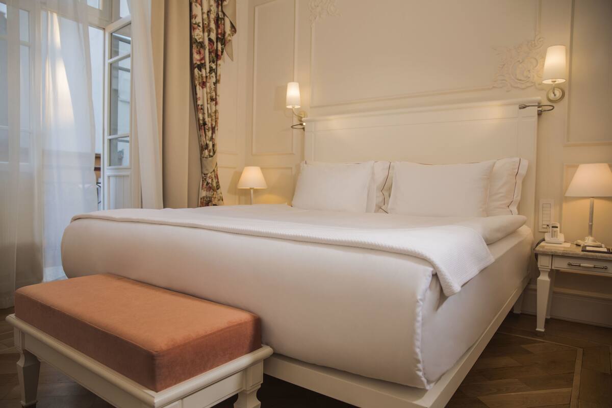 A neatly made bed with white linens, a matching bench, and two bedside tables with lamps.