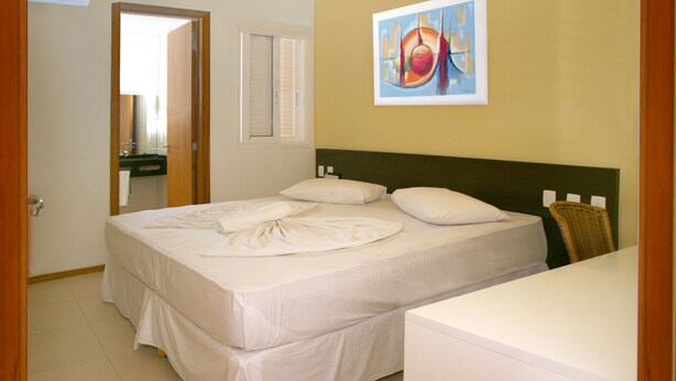 A hotel room with a bed, a bedside table, a chair, a painting on the wall, and a bathroom visible through an open door. - Saint Patrick Praia Hotel (Maceió)