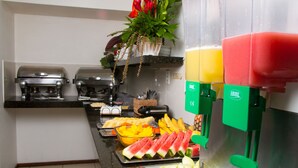 A buffet setup with various fruits, a coffee machine, and a juice dispenser. - Saint Patrick Praia Hotel (Maceió)