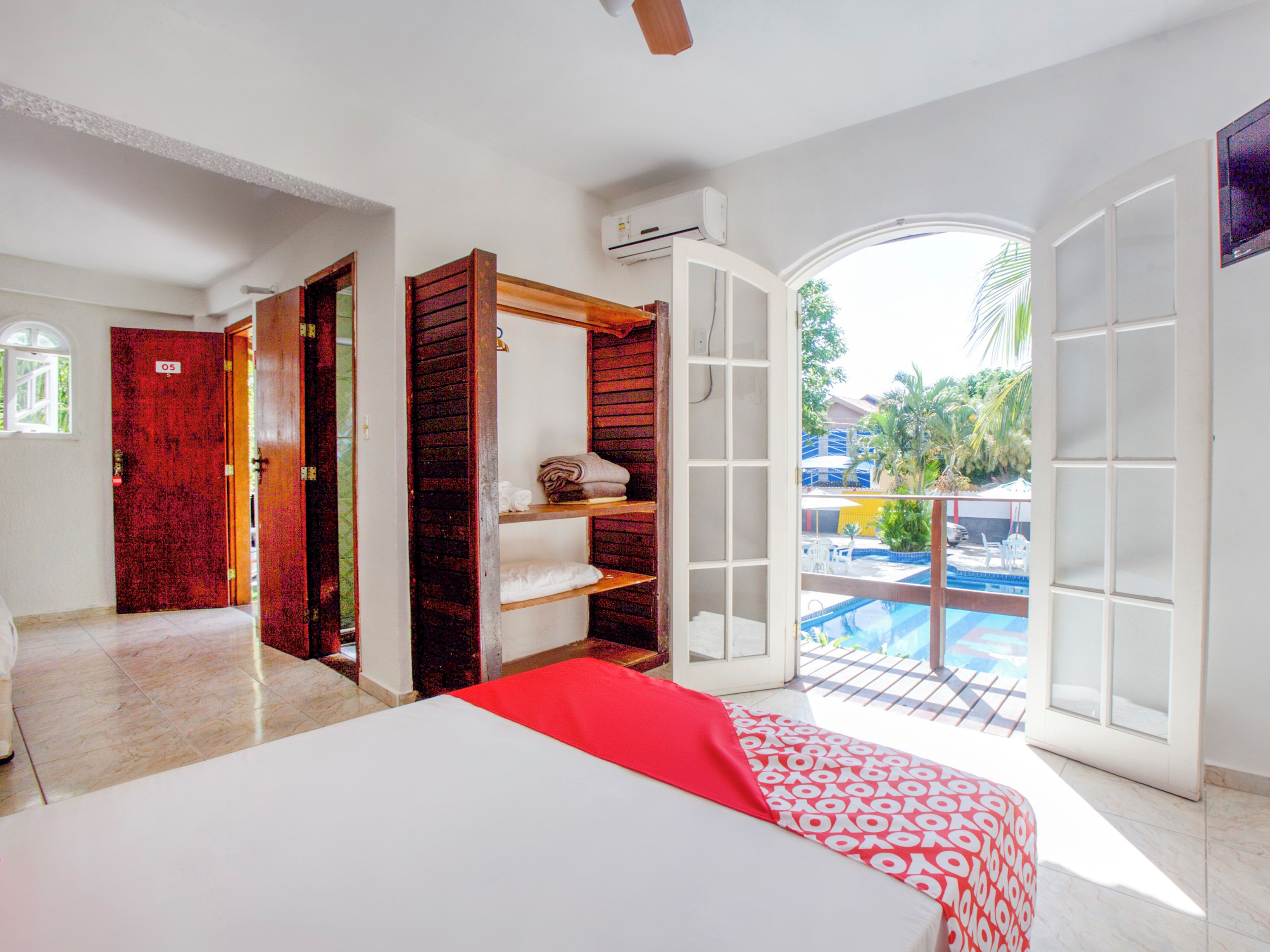 A hotel room with a bed, a red and white patterned bedspread, a wooden wardrobe, a white shelf, and a view of an outdoor swimming pool.