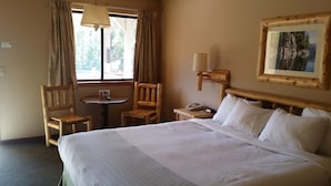 A hotel room with a large bed, two wooden chairs, a small round table, a lamp, and a painting on the wall. - Wildwood Inn (Twain Harte)