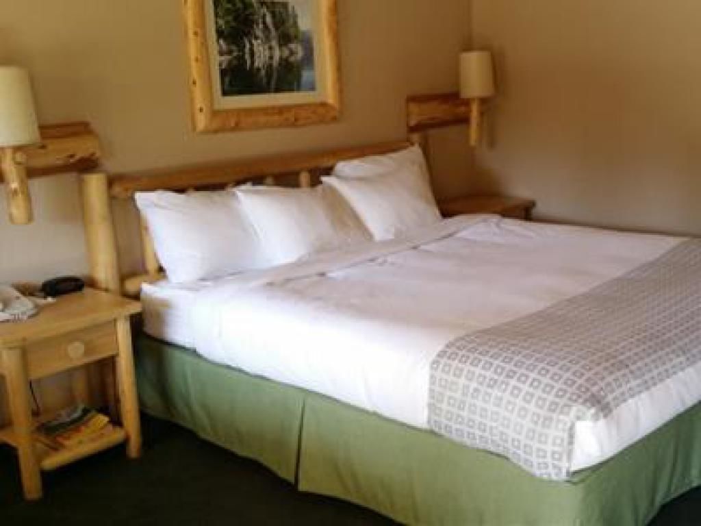 Cama bem arrumada com lençóis brancos e saia verde, duas mesas de cabeceira com abajures e um quadro na parede.