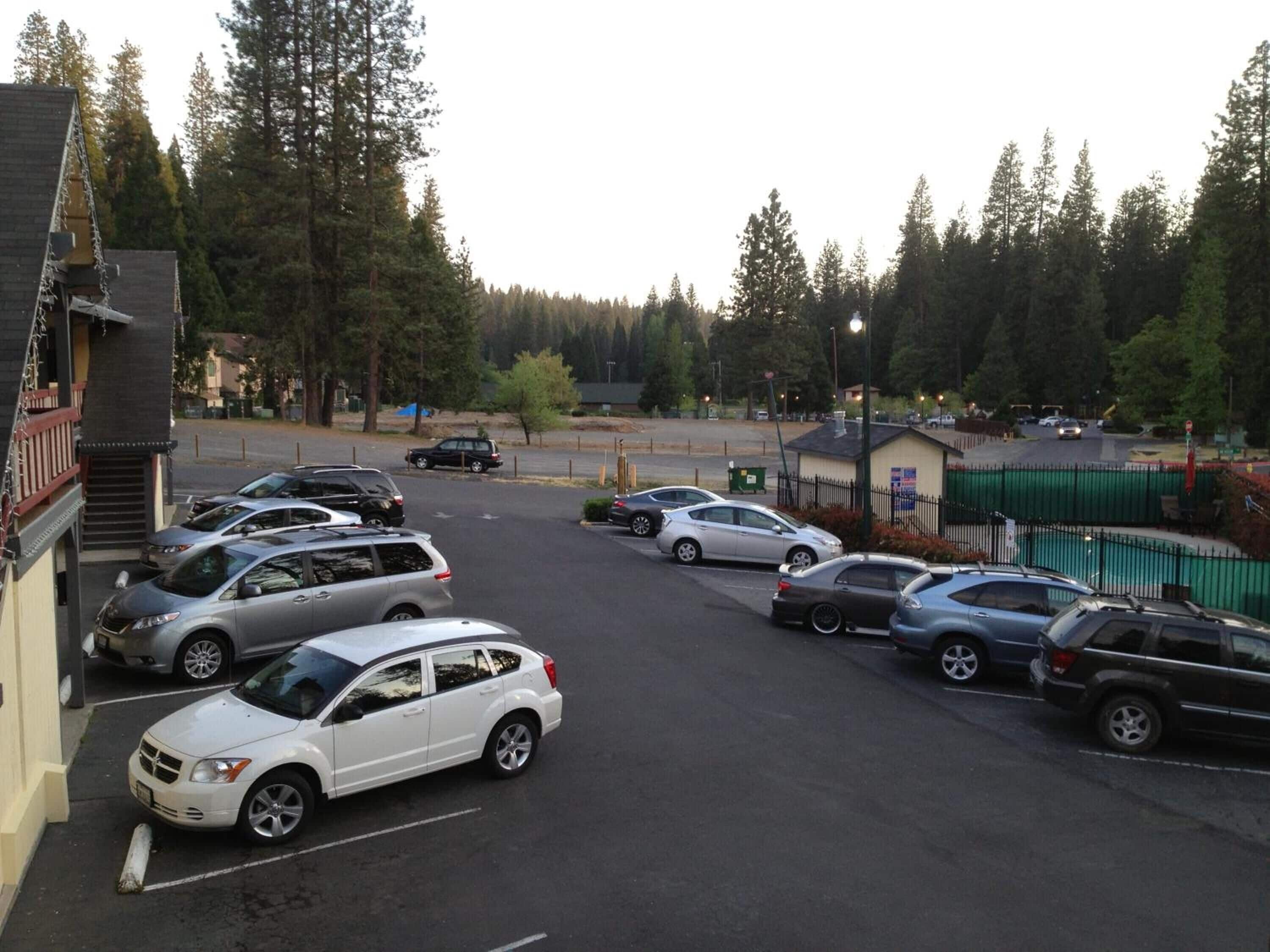 A parking lot with multiple cars, a building with a balcony, and a forested area in the background.